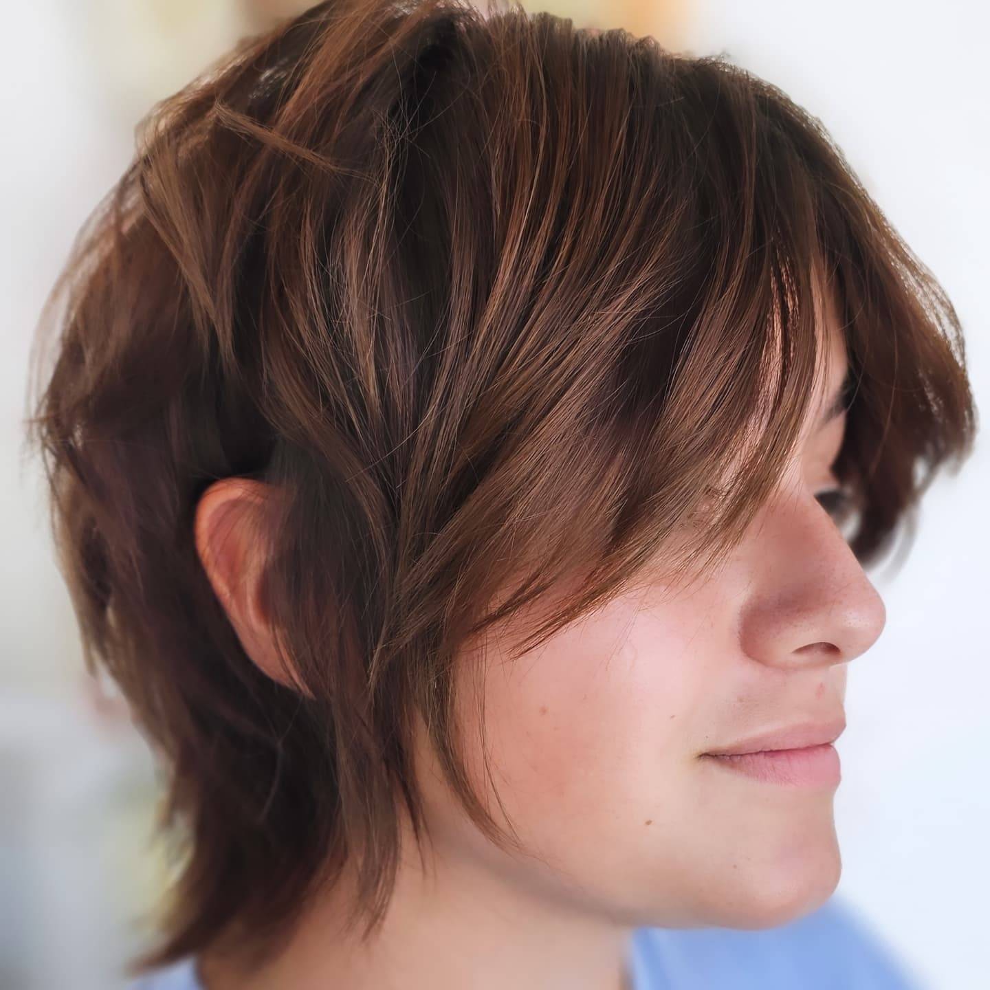 Side profile of a woman with short, layered brown hair and a soft, tousled style. She wears a light blue top.