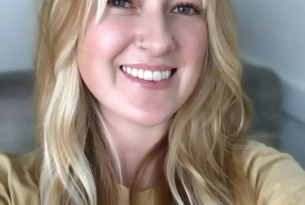 Close-up portrait of a smiling woman with long blonde hair and a nose ring, wearing a tan shirt.