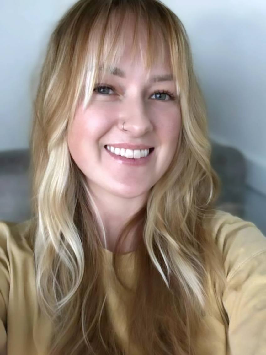 Close-up portrait of a smiling woman with long blonde hair and a nose ring, wearing a tan shirt.